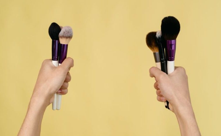 Contouring per principianti: tecniche semplici per definire il viso: Due mani che tengono vari pennelli per trucco su uno…